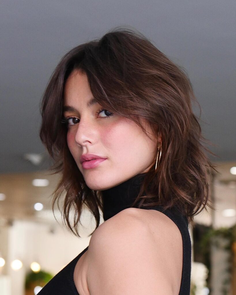 A woman looking over her shoulder showcasing a jaw length choppy mocha brown shaggy bob hairstyle with wispy curtain bangs and textured layers.