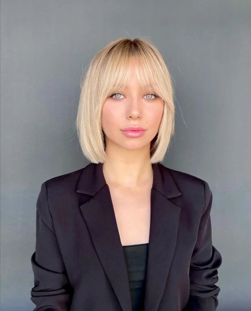 A young woman with green eyes looking forward, showcasing a straight champagne blonde angled bob haircut with wispy curtain bangs.