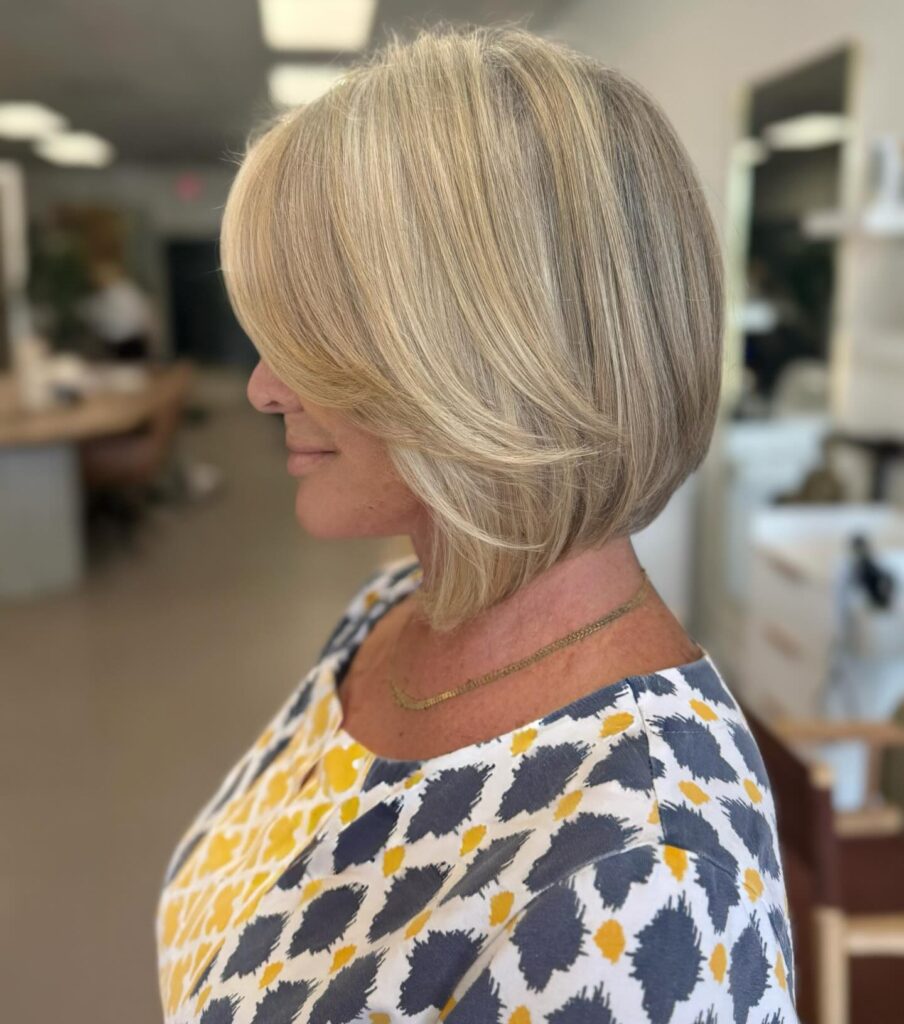 A side profile view of a woman showcasing a straight honey blonde chin-length graduated bob haircut with golden highlights and a side-swept front section.
