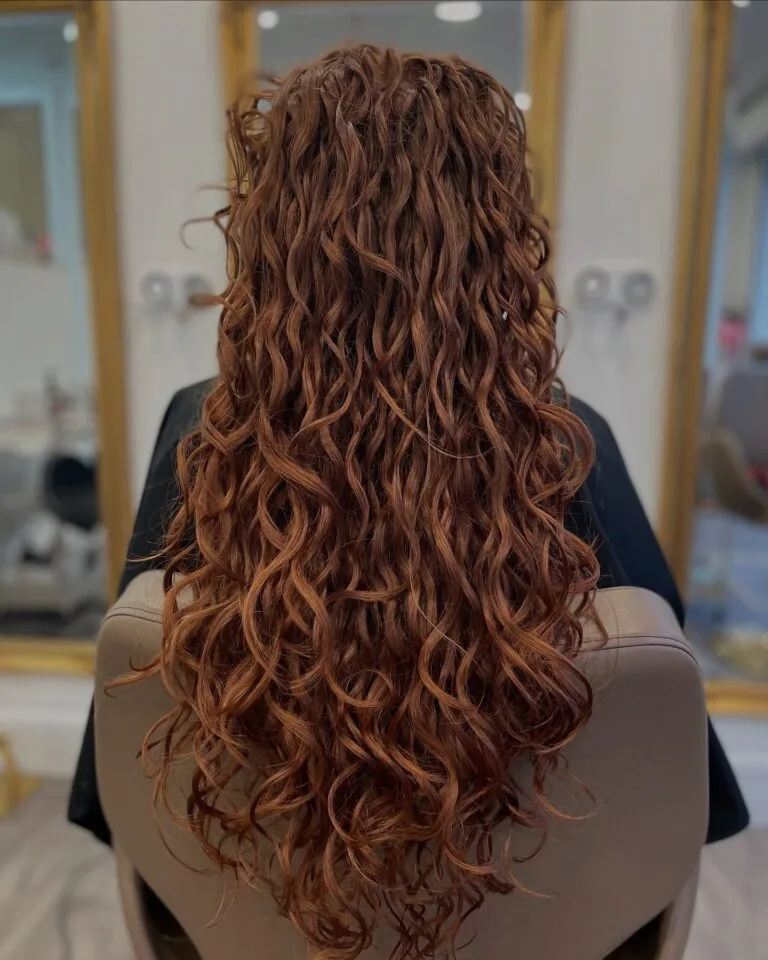 Long curly hairstyle with layered shaping and rich copper tones, featuring defined, cascading curls for full volume and movement.