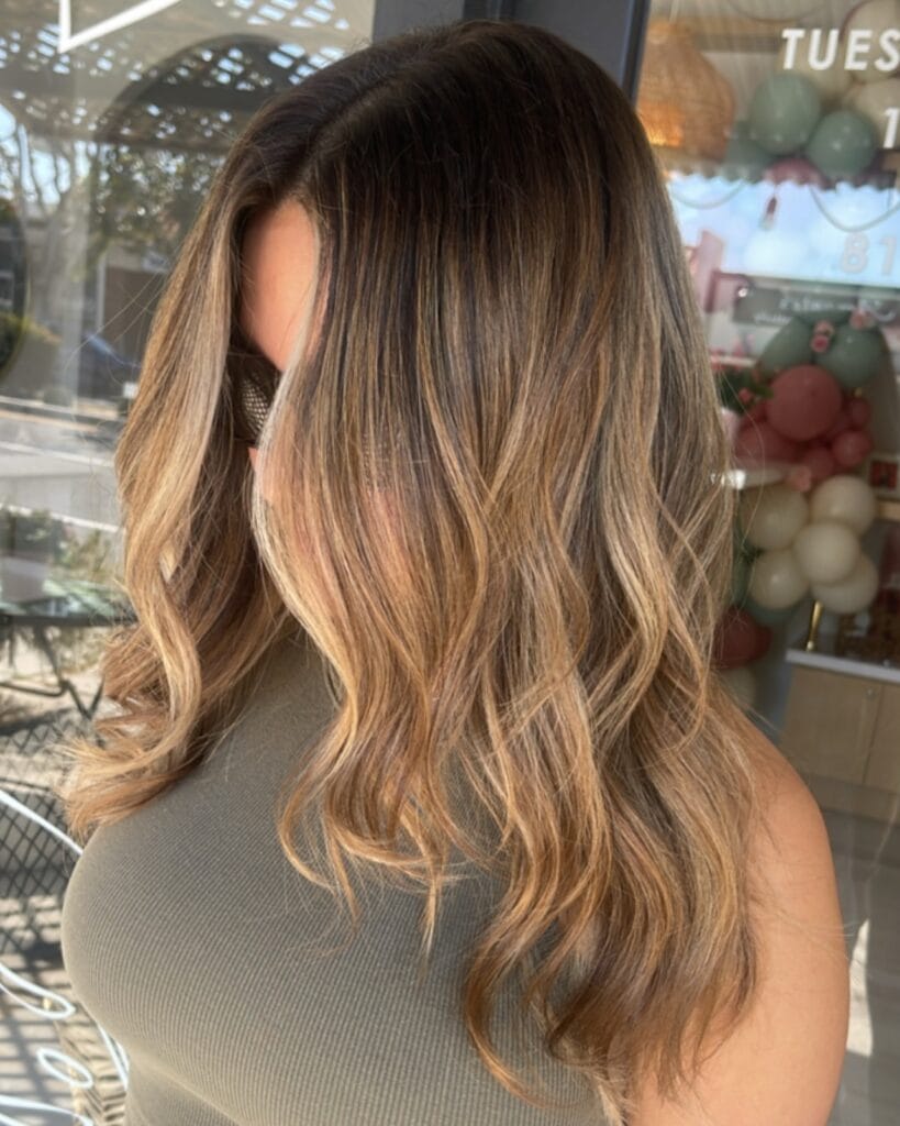 Medium-length wavy hairstyle with soft layers and warm honey brown balayage, blending darker roots into lighter golden ends for a natural, sun-kissed look.