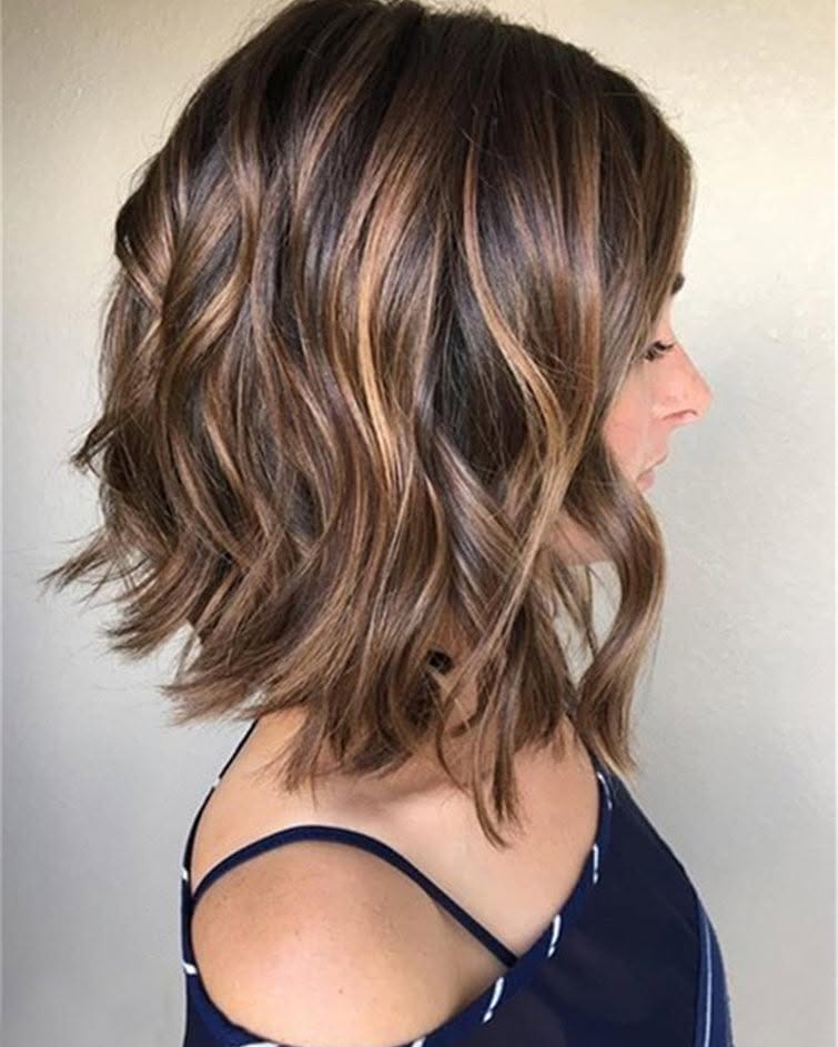 Shoulder-length wavy bob with soft layers and warm caramel balayage highlights blended through a medium brown base for a natural, dimensional look.