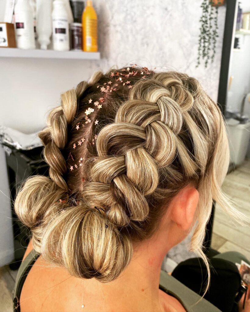 A woman with ash blonde hair styled in two thick Dutch braids ending in low buns with star shaped glitter on her scalp.
