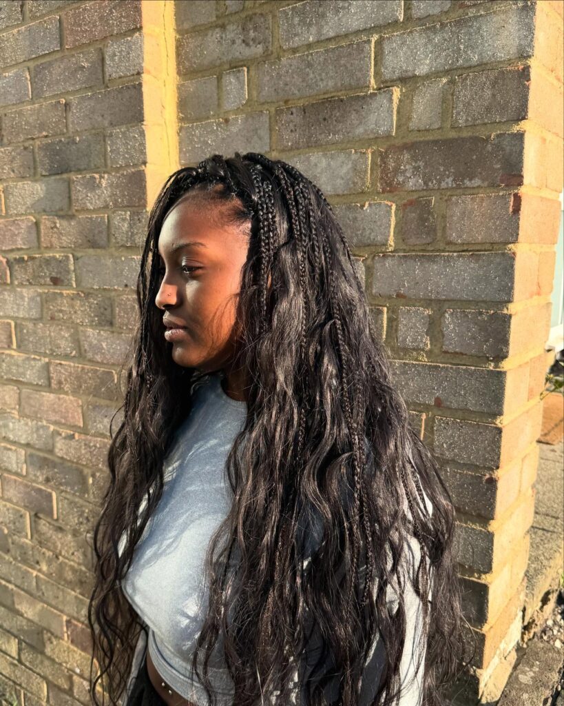 A young woman with long, dark wavy hair and a few thin knotless braids on the side looks profile in front of a brick wall.