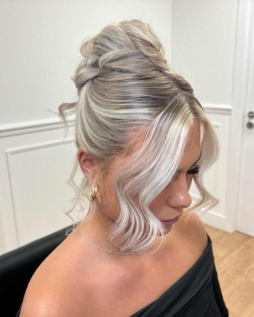 A young woman with highly textured and styled icy blonde hair featuring a large structural bun and wavy face framing pieces.