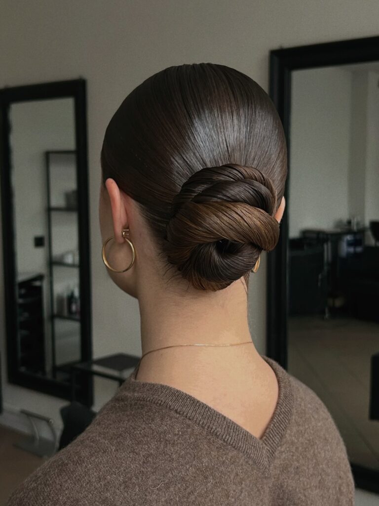 A back view of a woman with dark brown hair styled in a perfectly smooth twisted low bun.