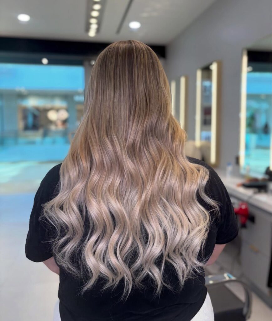 A woman with long hair featuring light brown roots that blend into platinum blonde waves, viewed from behind in a well-lit salon.