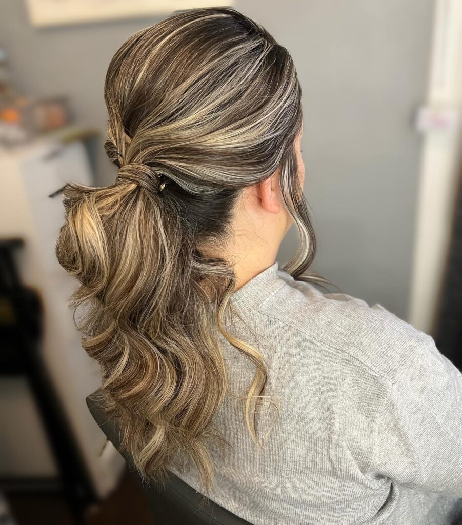 A woman from the side with dark hair and bright blonde highlights styled in a wavy low ponytail with a hair wrapped base.