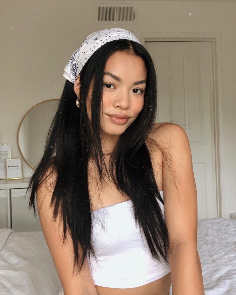 A woman with long straight black hair wears a white patterned bandana tied over her head in a kerchief style.