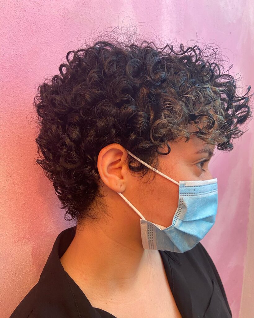 A side profile of a woman with a dark, short, curly pixie cut featuring subtle brown highlights and a tapered back.