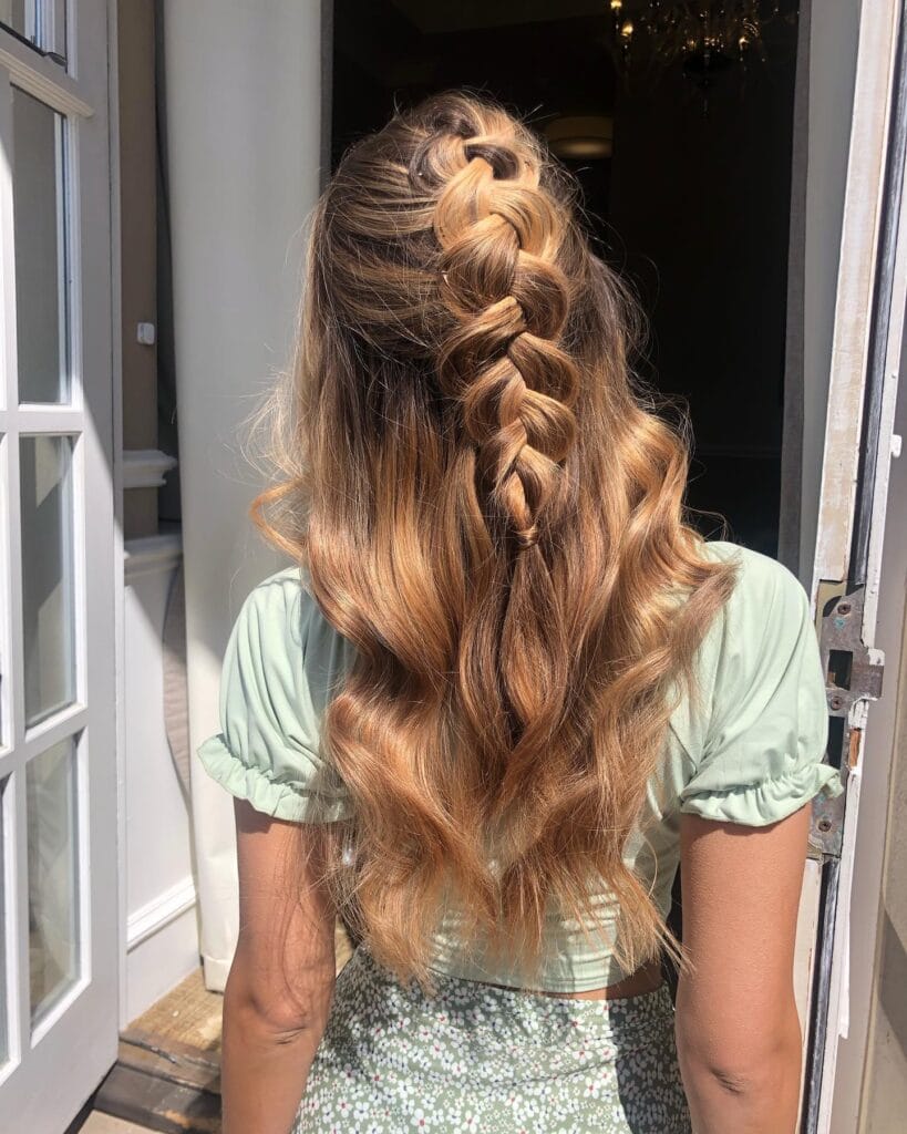 A woman with long wavy brown hair with honey blonde highlights has a central French braid in a half up style with flowing loose waves below.