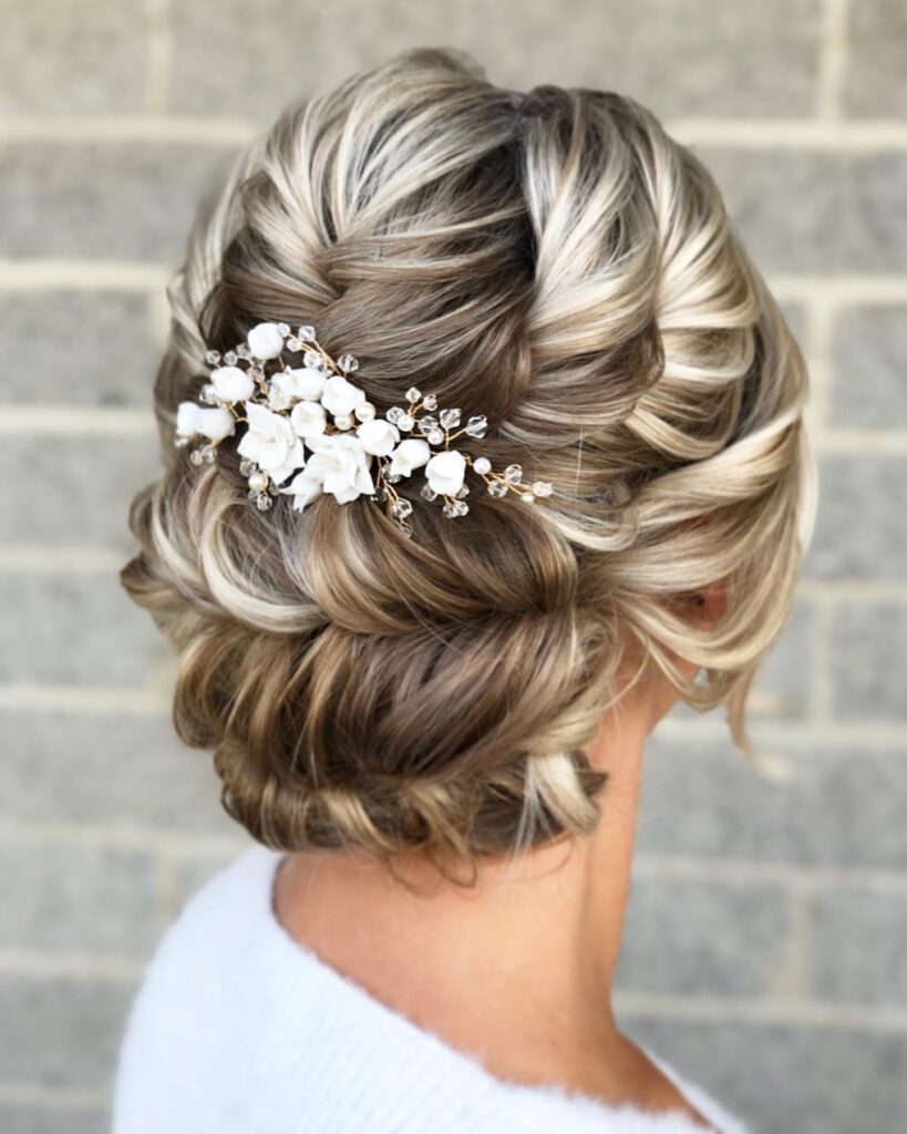 A woman with dimensional ash blonde hair styled in a complex braided low updo adorned with a white floral hair accessory.