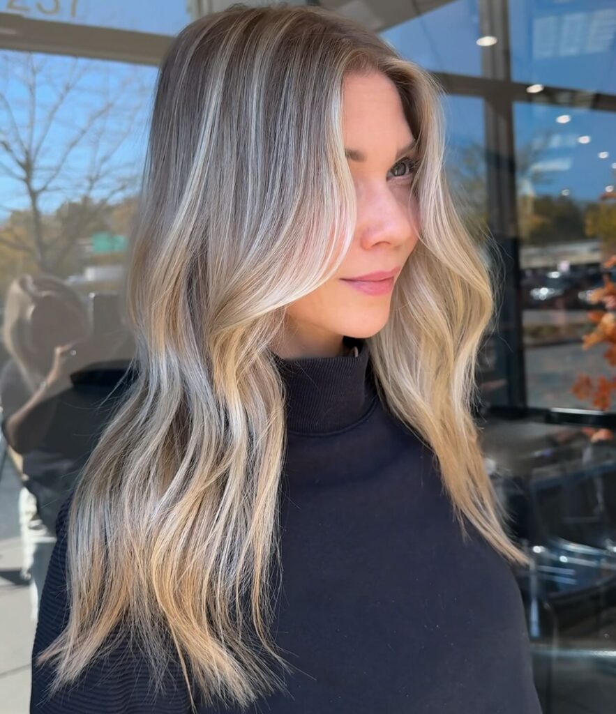 A woman with long ash and sandy blonde hair styled in a straight and wavy mix with prominent face-framing money pieces.