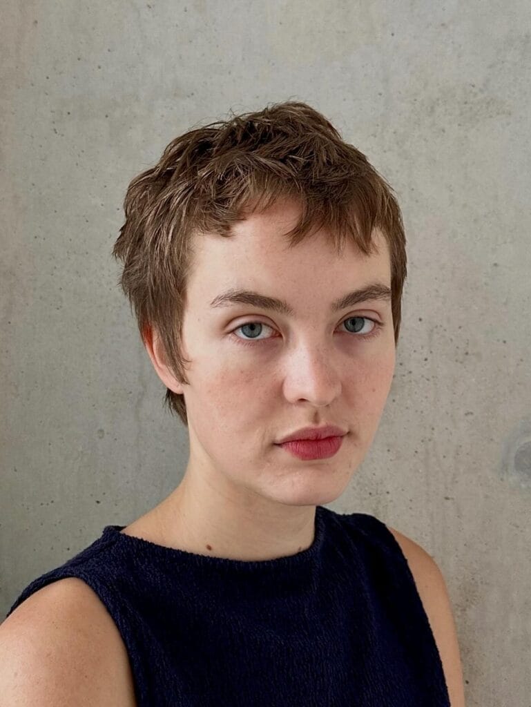 A woman with a short choppy micro pixie haircut in a natural light brown shade posing against a grey wall.