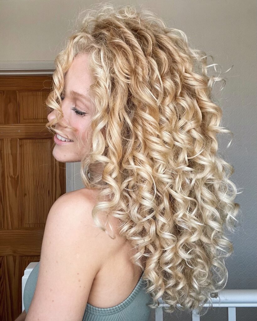 A woman with long cascading champagne blonde curls showing off her voluminous and highly defined ringlets from a side profile.