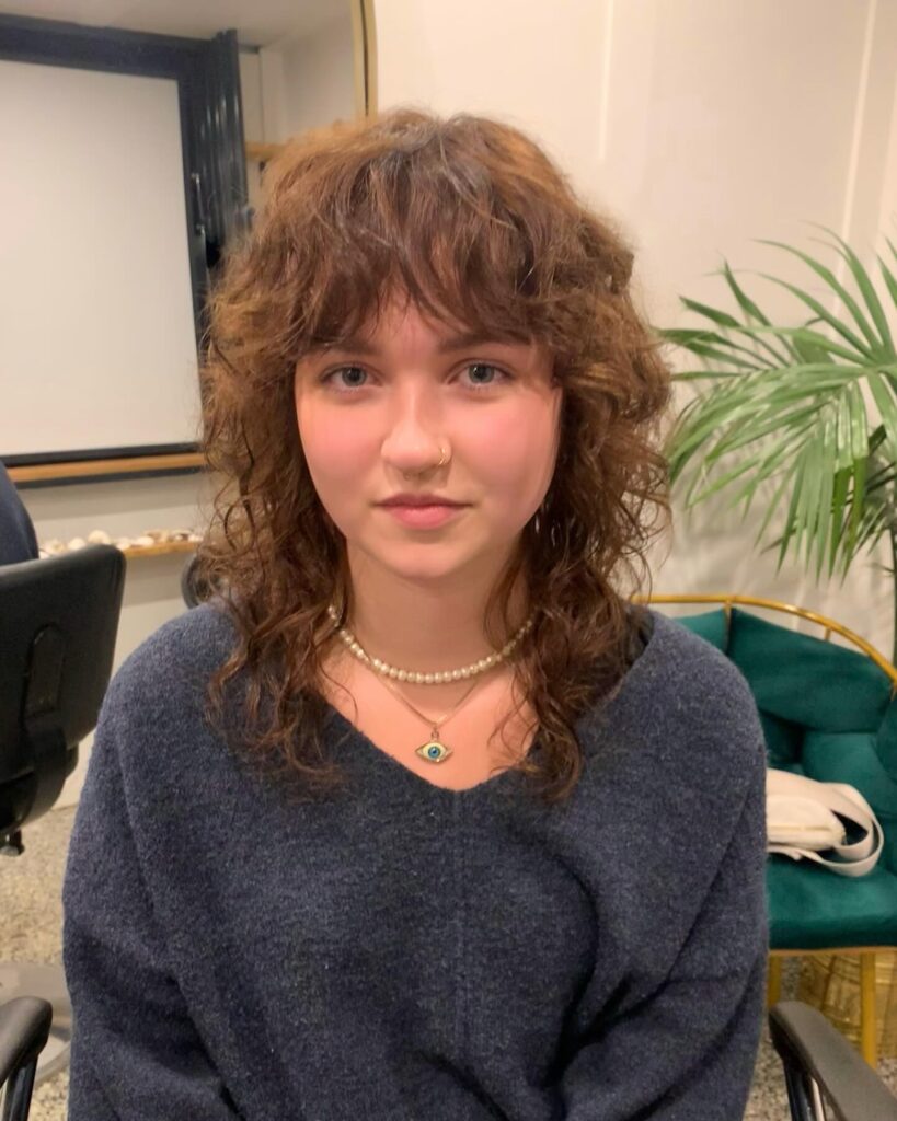 A young woman with curly light brown hair in a heavily layered shag haircut with a textured curly fringe.