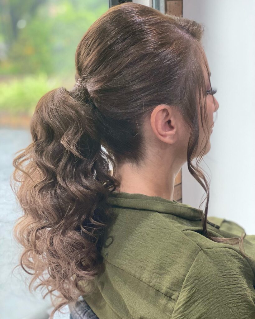 A side profile of a woman with ash brown hair in a mid height ponytail featuring voluminous beachy curls and loose face framing tendrils.