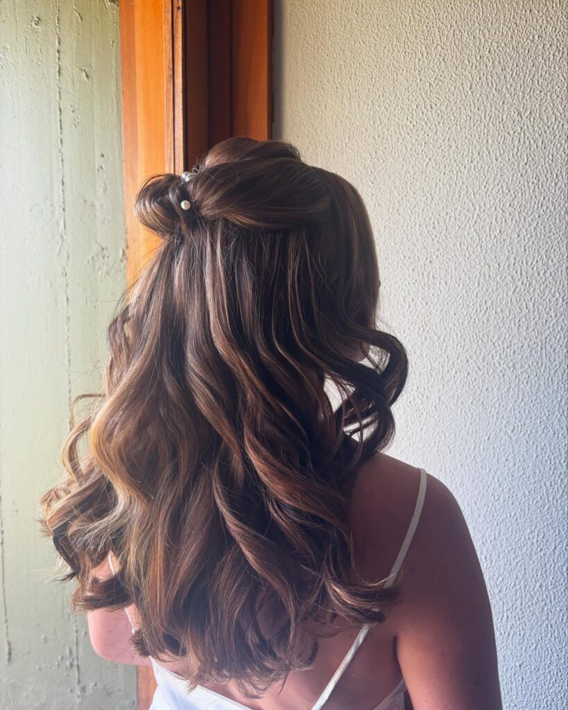A rear view of a woman with long, wavy, brown hair styled in a half-up twisted bun decorated with small pearl hairpins.