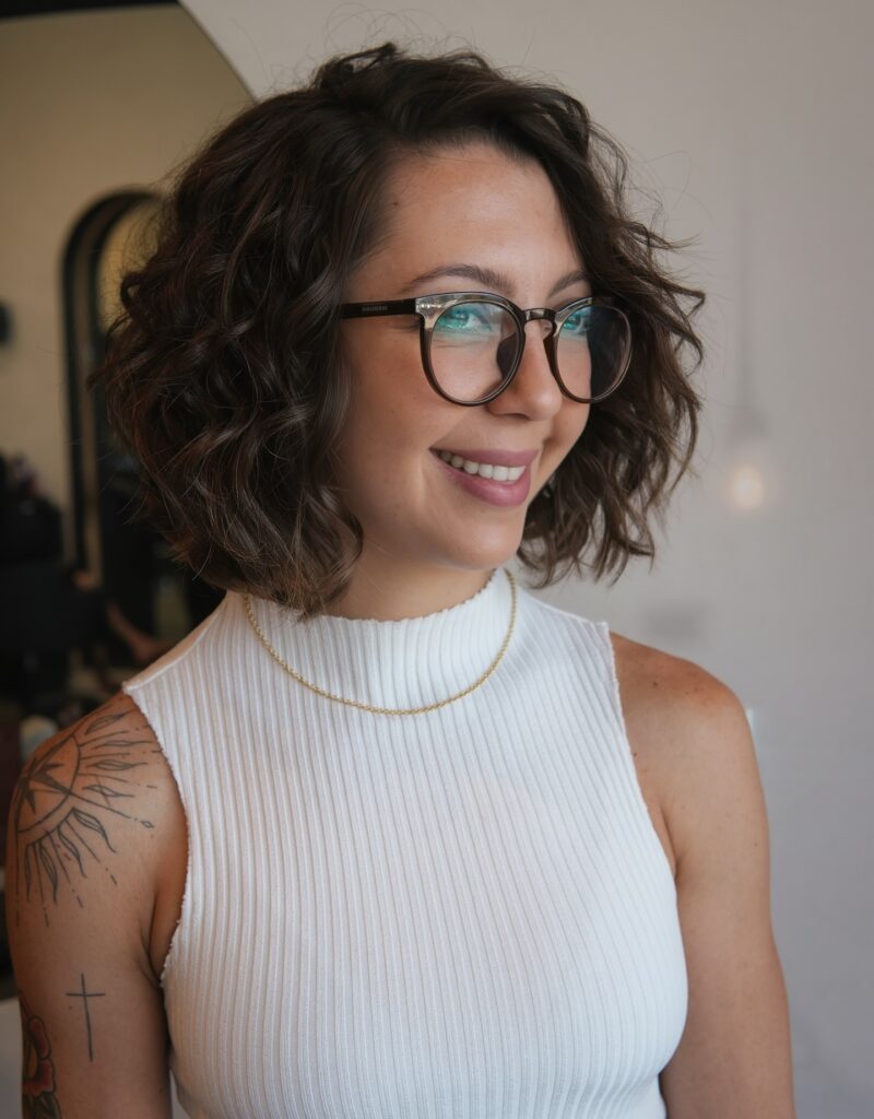 A smiling woman wearing glasses with a brunette wavy lob that features subtle highlights and voluminous texture.