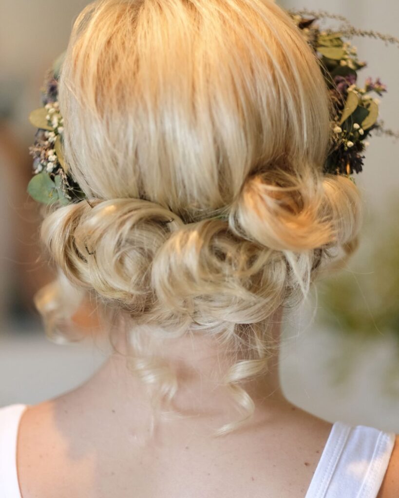 A woman from behind with platinum blonde hair styled in soft pin curls and adorned with a full crown of green eucalyptus leaves and tiny flowers.
