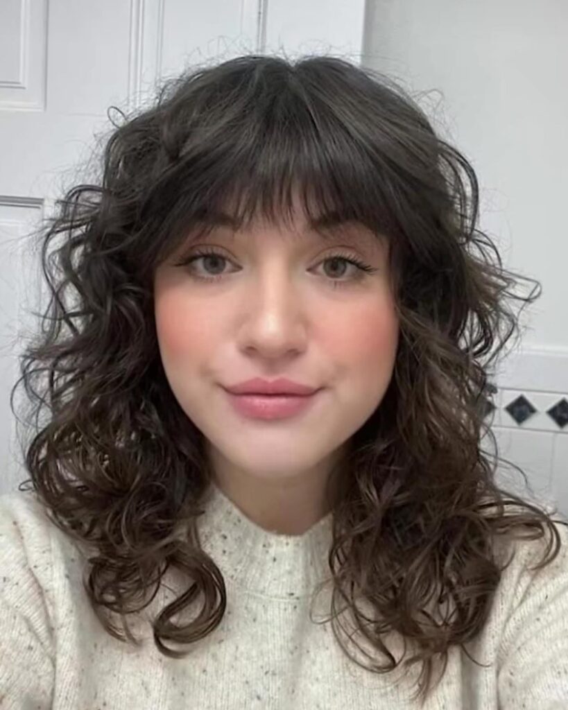 A woman with medium-length dark brown wavy hair featuring a full, wispy fringe.