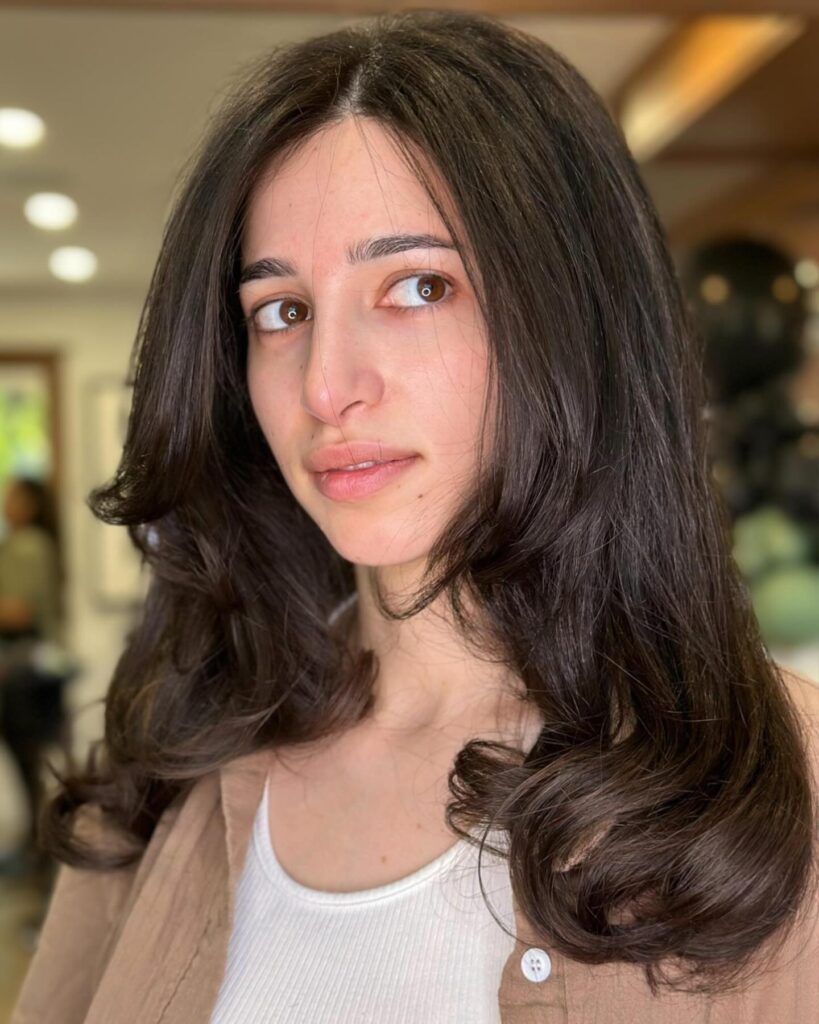 A woman with voluminous medium length espresso brown hair featuring bouncy layers and face framing curtain bangs.
