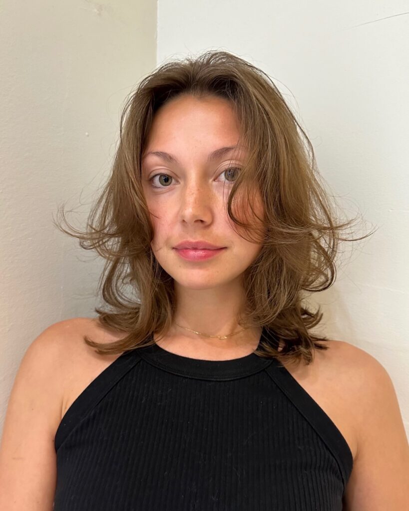 A woman with shoulder length ash brown hair featuring wispy textured layers and face framing flicks.