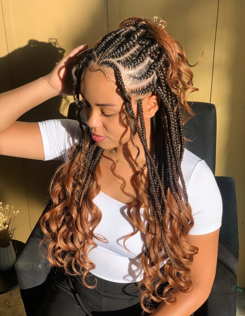 A woman with copper toned Fulani braids styled in a half up ponytail with large bouncy curls at the ends.
