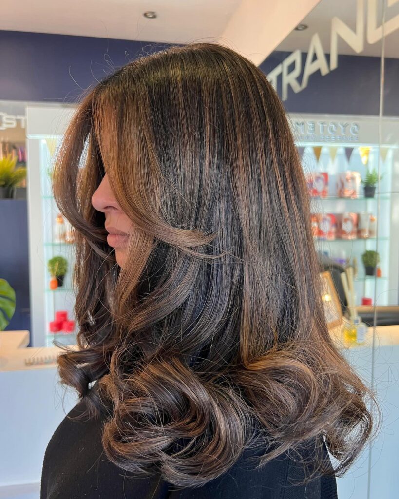 A woman with medium-long toffee brown hair featuring face-framing layers and large bouncy curls at the ends.