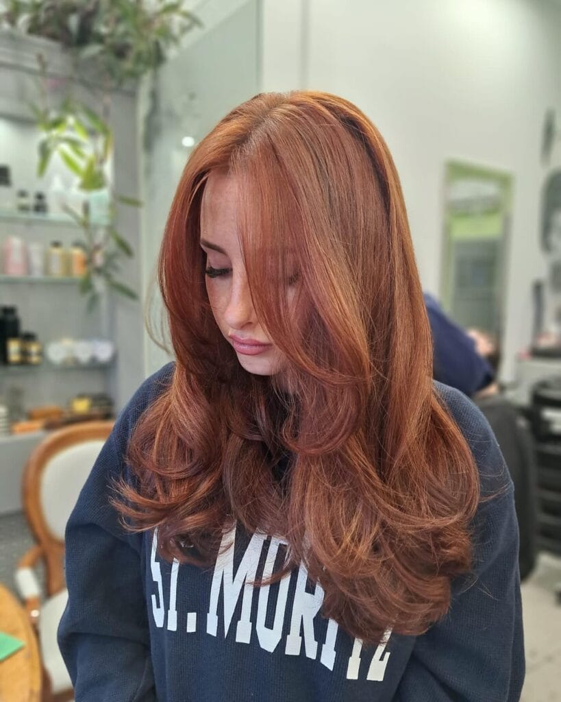 A woman with long vibrant copper red hair styled with voluminous face framing layers and curled ends.