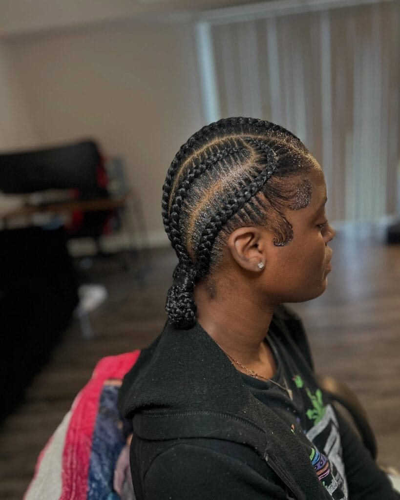 A woman shows a side view of her dark cornrow braids that curve into a low bun with detailed swirl designs on her edges.