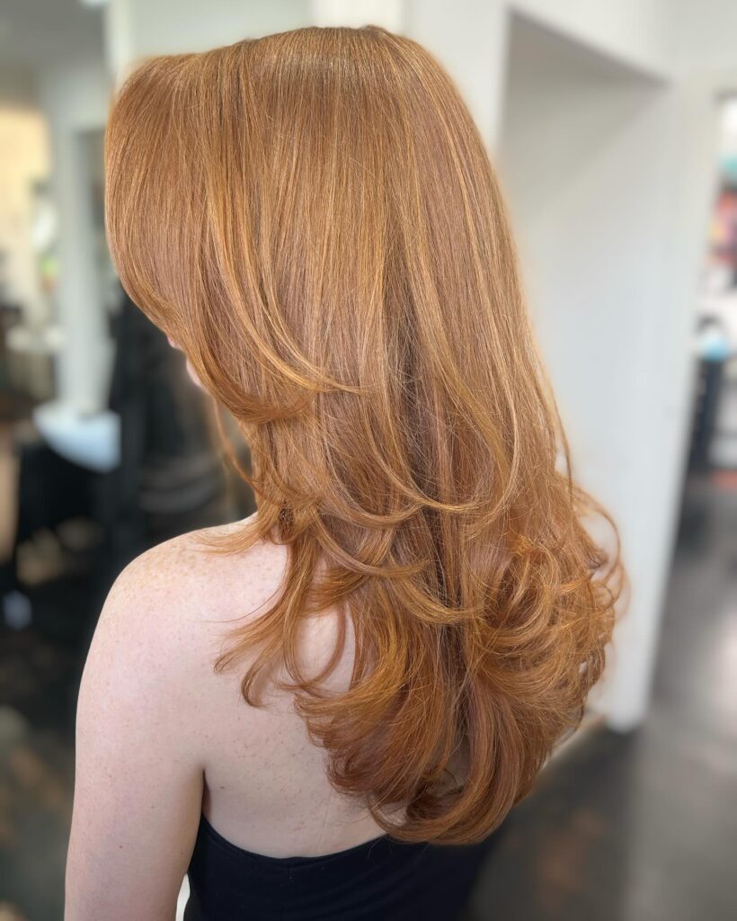 A back view of a woman with long red copper hair styled with face framing layers and bouncy outward curled ends.