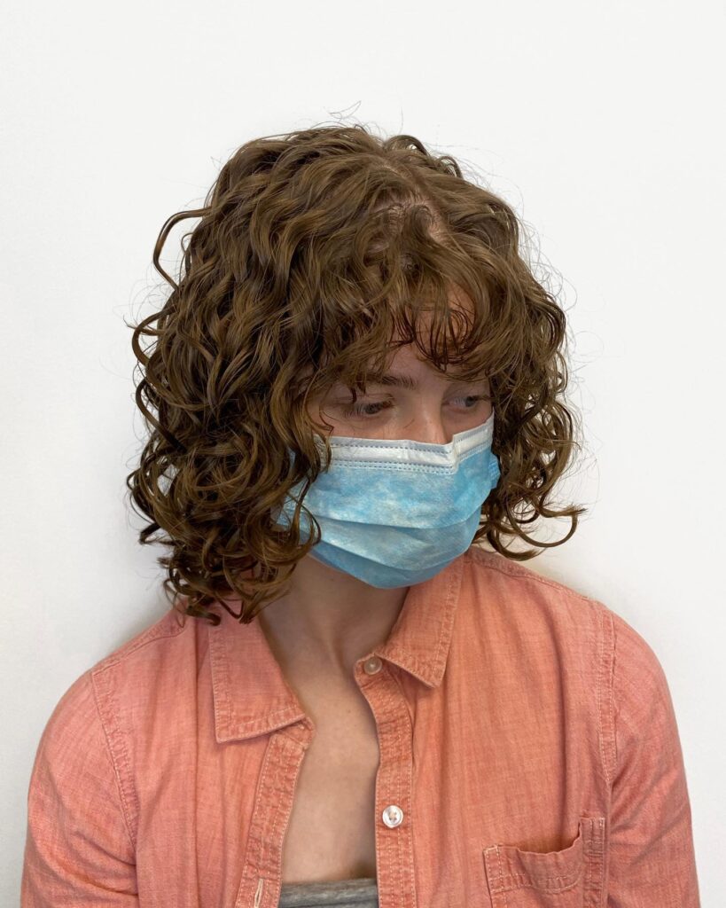 A woman with defined shoulder length curls and full fringe in medium brown tones wearing a mask.