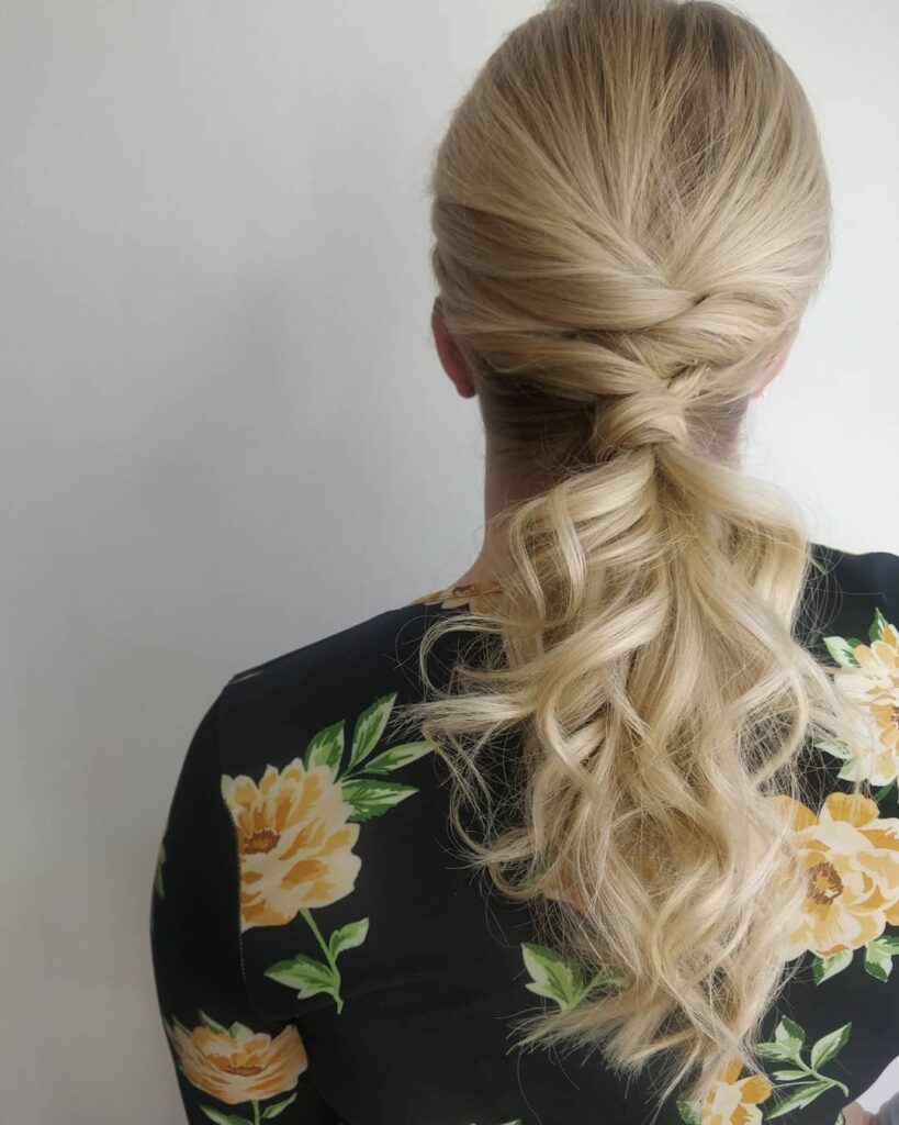 Long blonde hair styled in a low twisted ponytail with soft voluminous curls.
