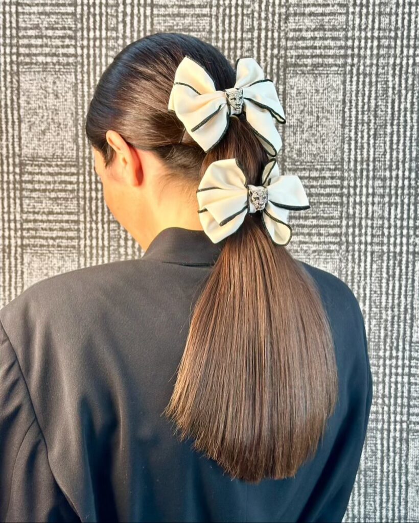 A woman with a sleek brown ponytail wears two stacked cream bows with black trim down the length of her hair.