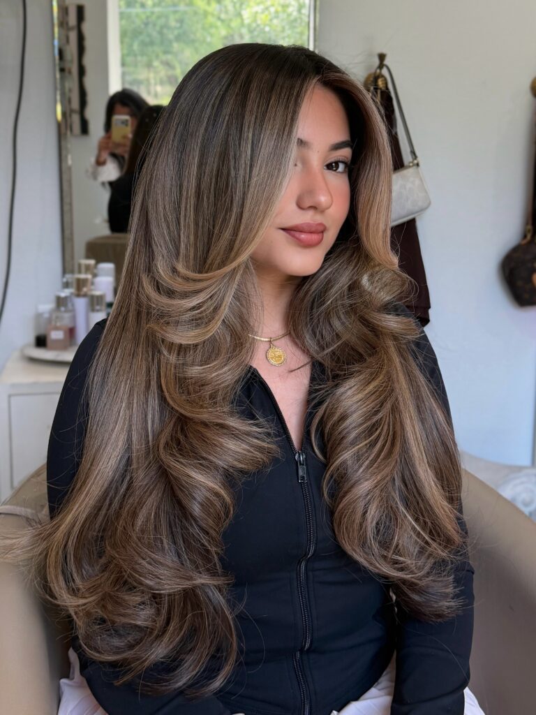 A woman with long bronde balayage hair styled in bouncy waves and swept back curtain bangs wearing a black outfit.