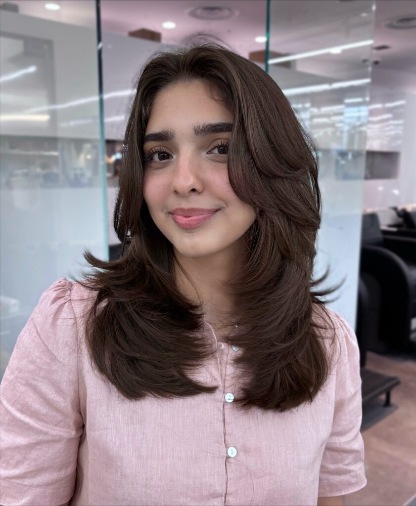 A woman with medium length brown hair styled in flipped out layers and soft curtain bangs wearing a pink top.