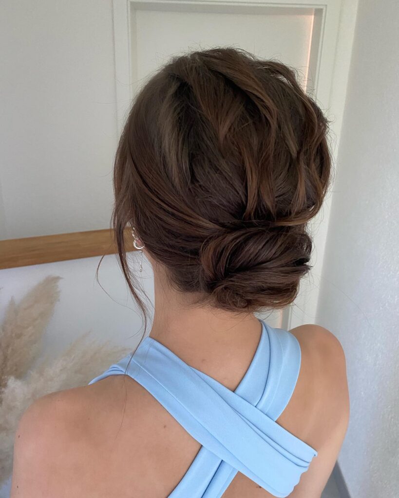 A woman with wavy brunette hair styled into a textured low bun with soft face-framing pieces.