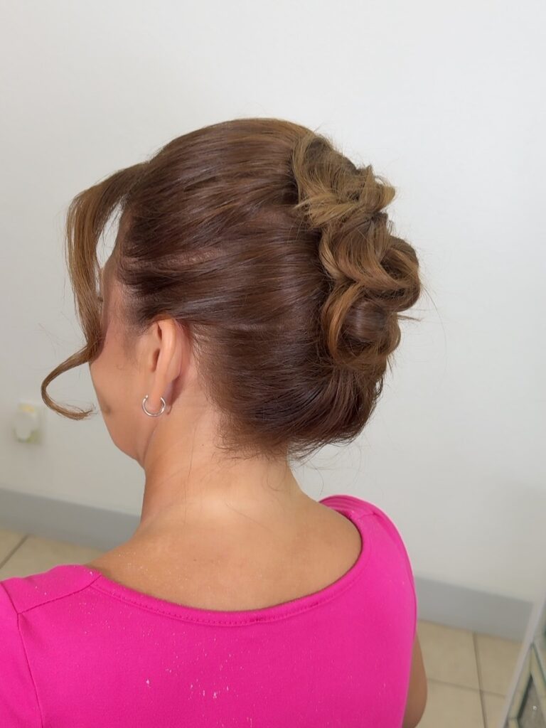A woman with brown hair featuring caramel highlights wears a sleek low curled bun with loose curled strands framing her face.