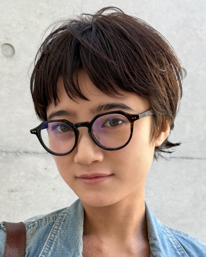 A woman wears a textured brunette pixie cut with choppy layers and soft fringe, paired with round glasses.