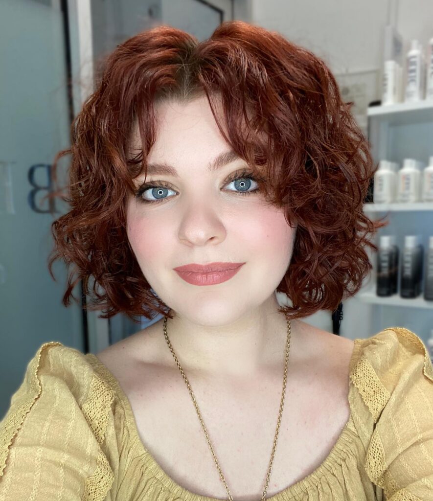 A woman with a chin-length curly copper red bob styled with a center-parted curtain fringe.