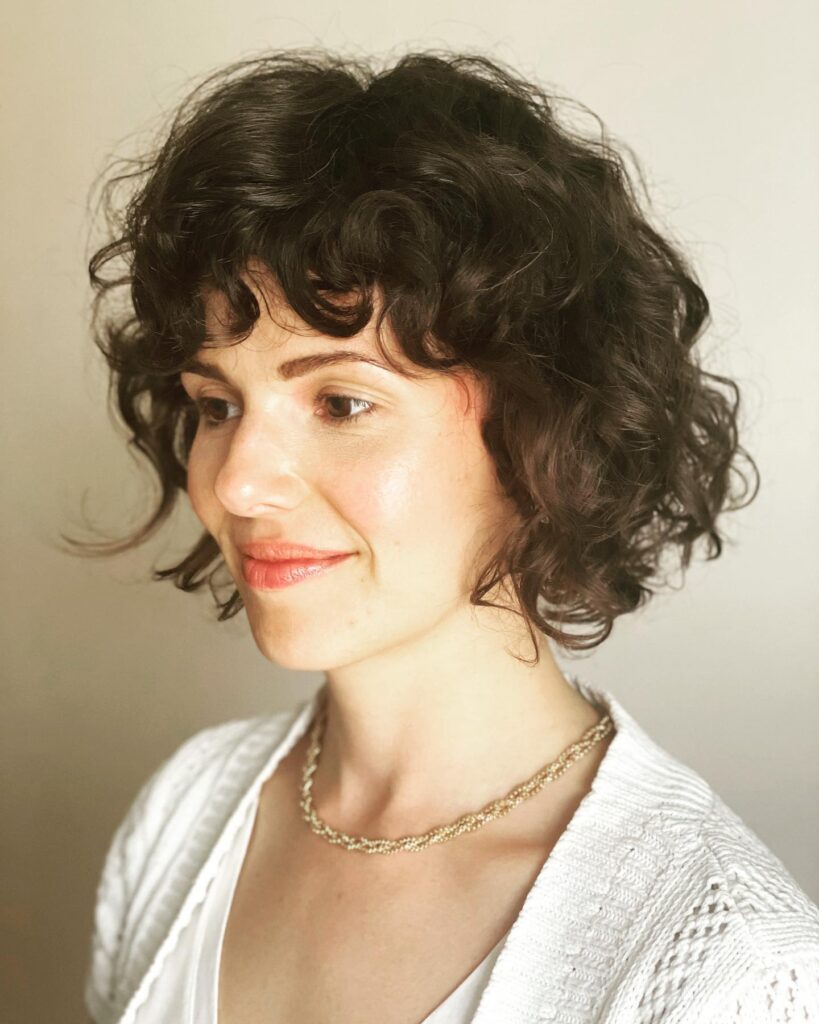 A woman with a jaw-length dark brunette curly bob styled with soft wispy center-parted bangs.