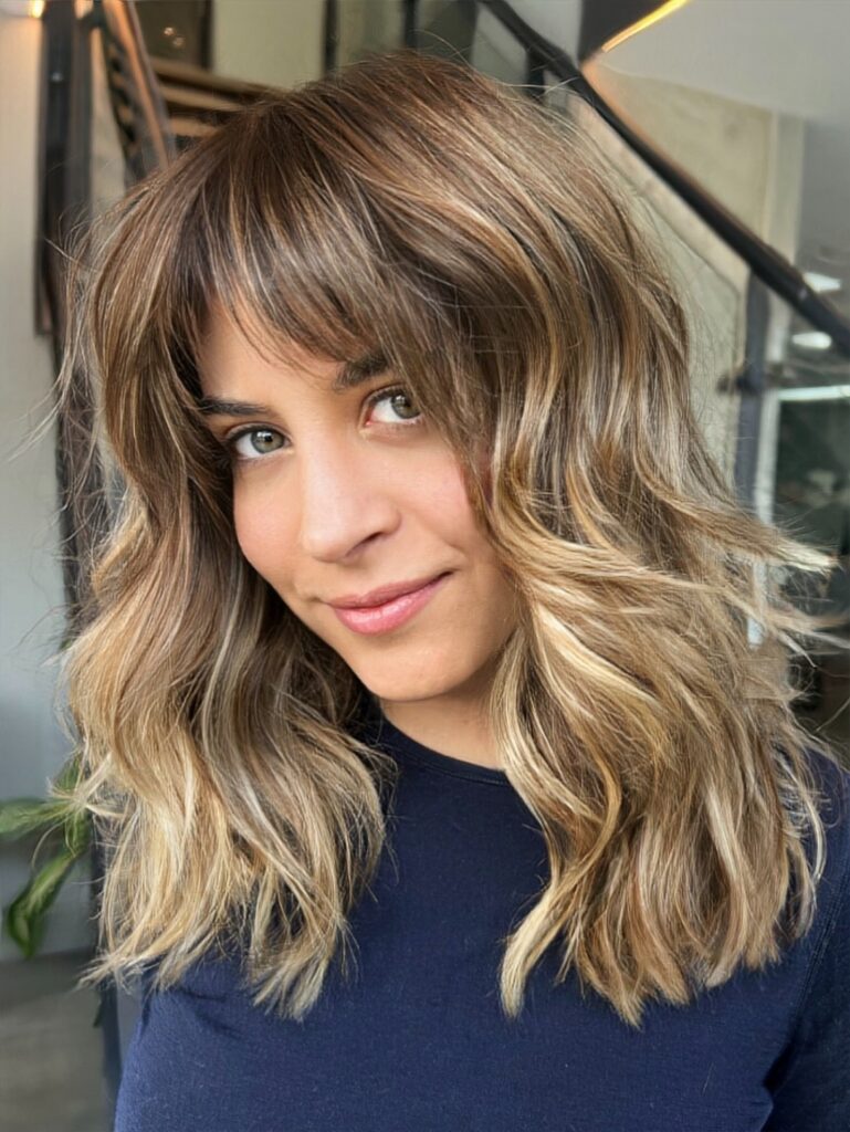 A woman with medium length brunette to blonde balayage hair featuring wispy bangs and soft waves.