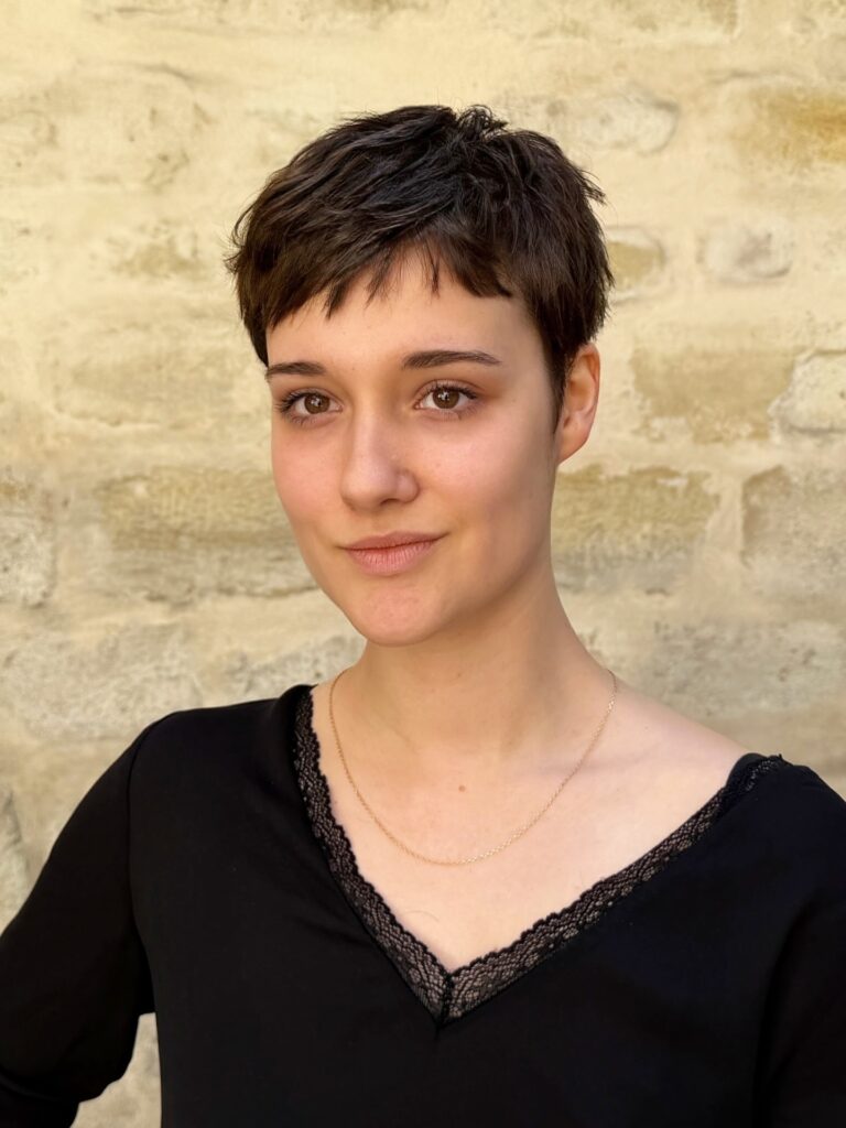 A woman with a short textured dark brunette pixie cut and side swept fringe poses against a stone wall.