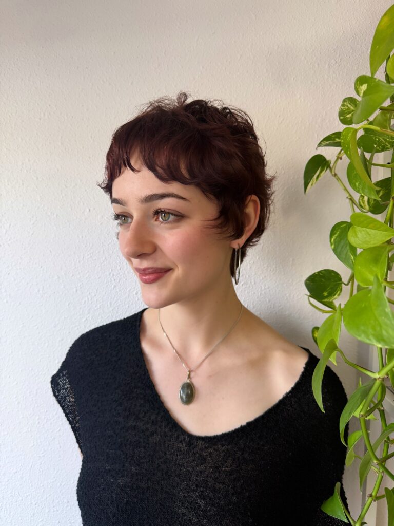 A woman with a curly deep auburn pixie cut and wispy fringe smiles while wearing a pendant necklace.