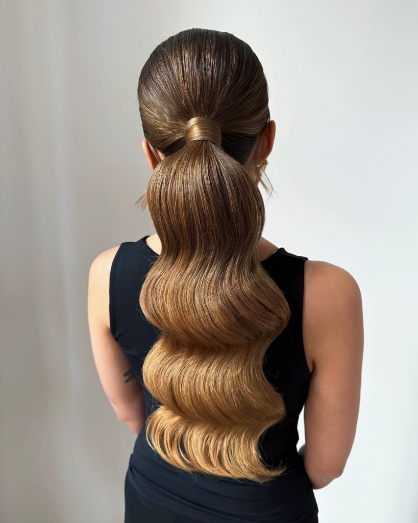 A woman with caramel brown hair wears a voluminous low ponytail with a bumped crown and glossy Hollywood waves.