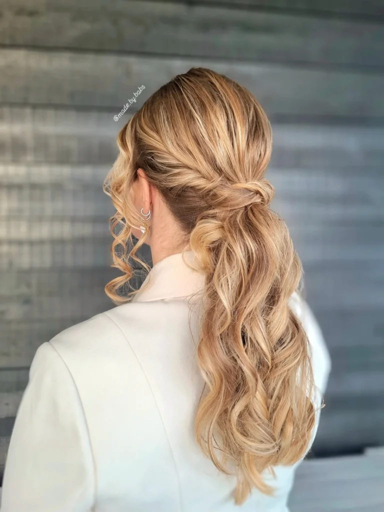 A woman viewed from behind wears a low honey blonde ponytail with twisted sides and bouncy beachy curls.