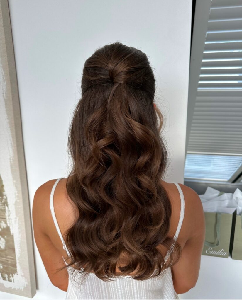 A woman with long brunette hair styled in a sleek half up twist with cascading curls down her back.