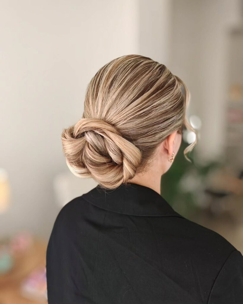 A woman with highlighted blonde hair styled in a low woven knot with delicate wavy tendrils around her face, seen from behind wearing a black blazer.