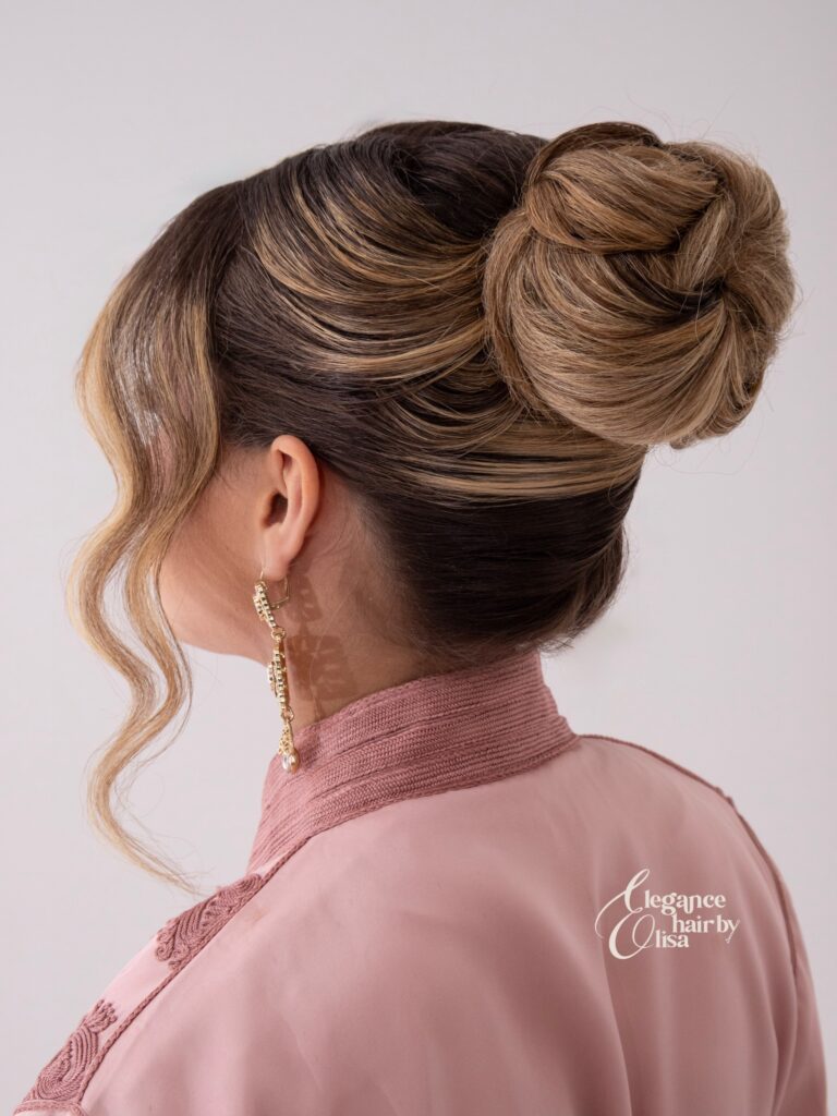 A woman with brown and blonde balayage hair styled into a sleek high wrapped topknot with wavy face framing strands, shown in profile wearing a pink outfit